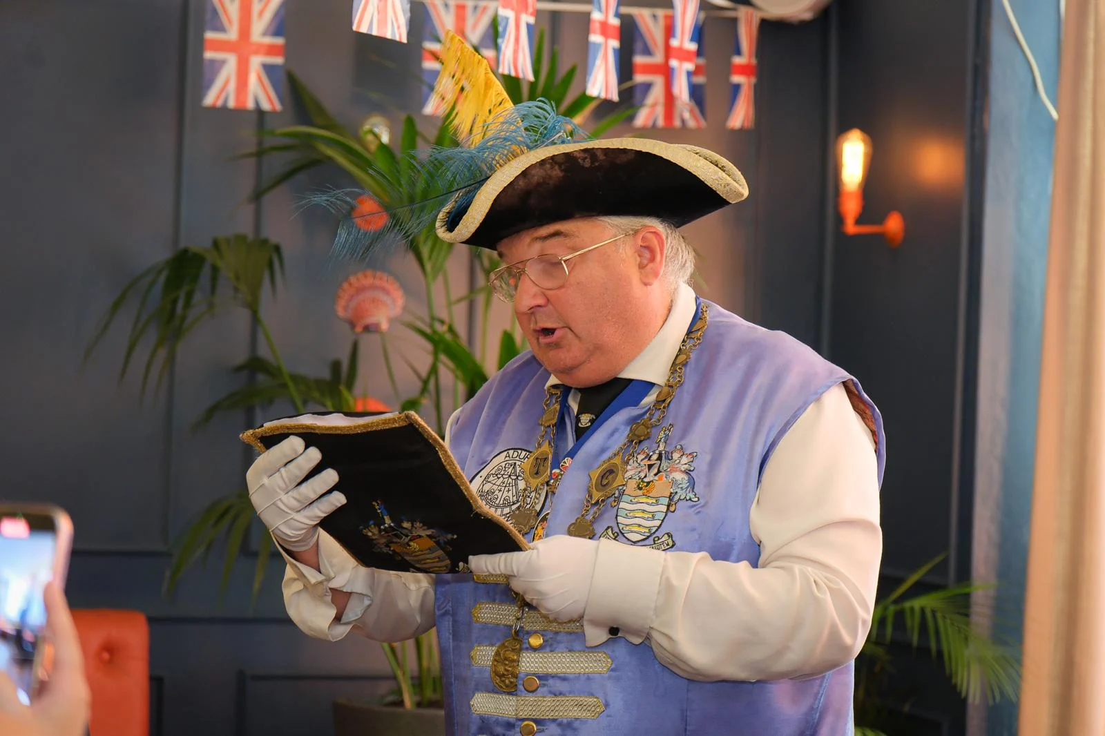 Bob Smytherman reading a proclamation from his ceremonial book at an indoor business event, beneath Union Jack bunting.